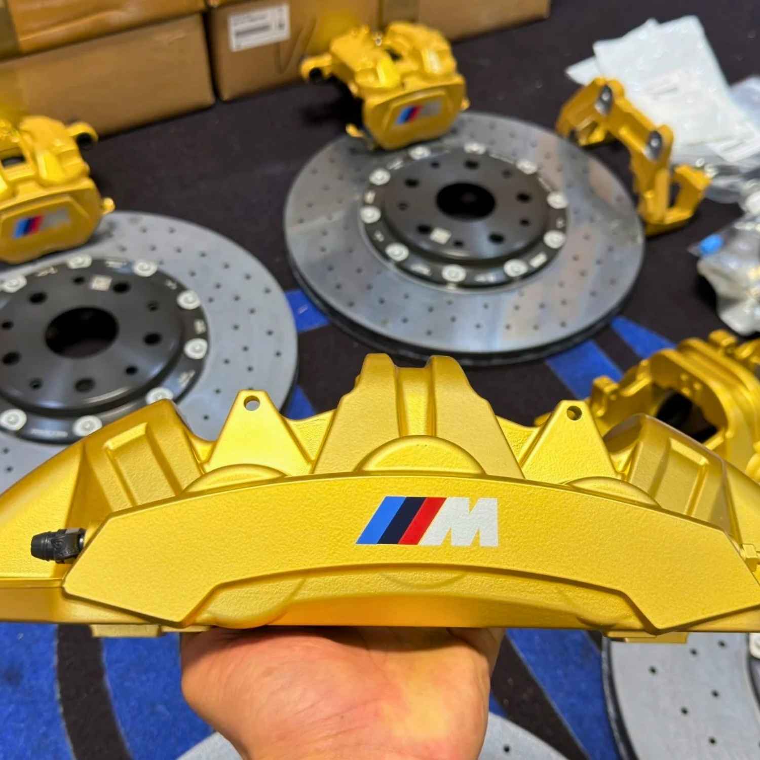 Hand holding a gold BMW M Carbon Ceramic brake caliper, shown with a complete set of ForzaCCB carbon ceramic brake rotors.