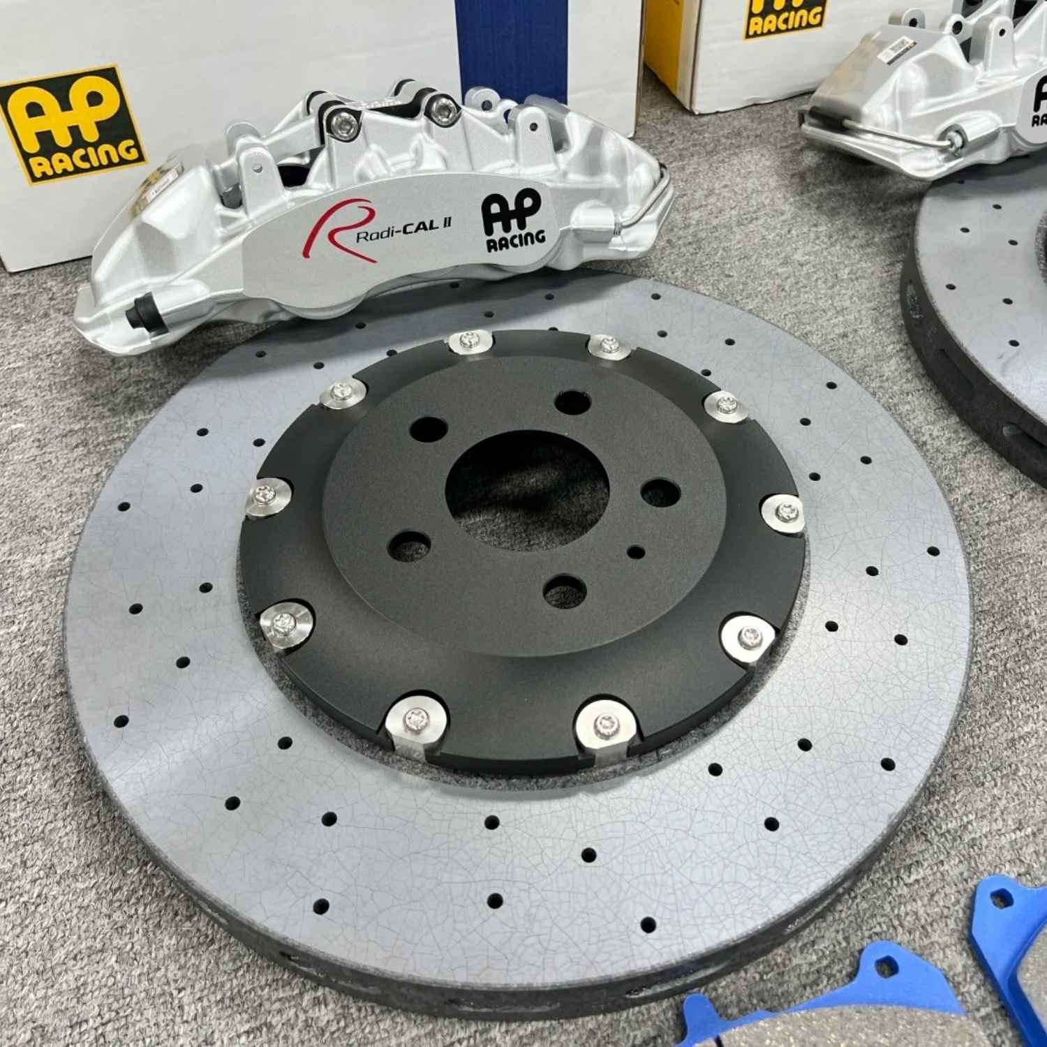 High-performance brake upgrade kit featuring a ForzaCCB carbon ceramic rotor and silver AP Racing Radi-CAL II caliper with AP Racing packaging in the background.