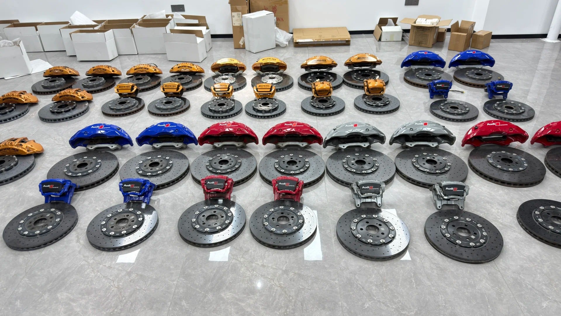 Large collection of genuine Audi and Mercedes-Benz performance brake components arranged on a showroom floor, including drilled rotors and blue, red, gray Audi calipers alongside bronze AMG calipers for carbon ceramic and big brake system upgrades.